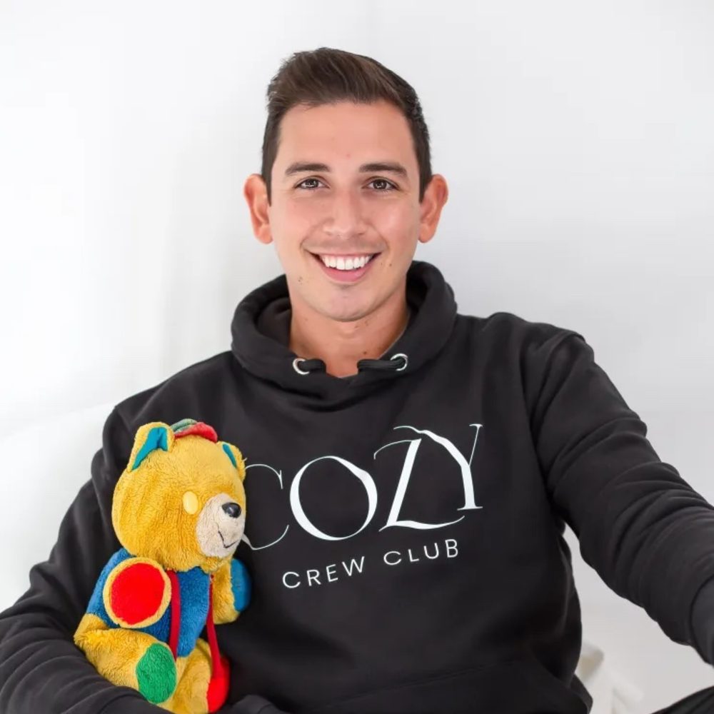 Conor Jaafari, founder of Cozy Crew Club, smiling holding an old multi-coloured teddy bear 