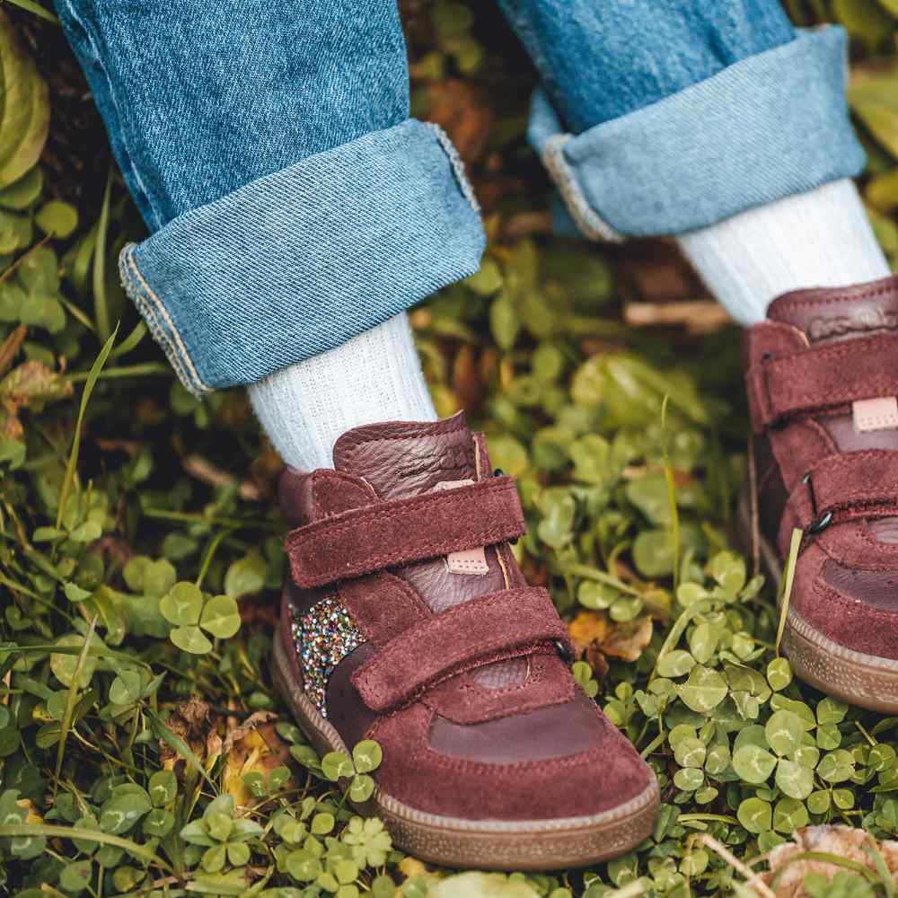 A close up of a child's feet on grass wearing maroon coloured shoes by Froddo