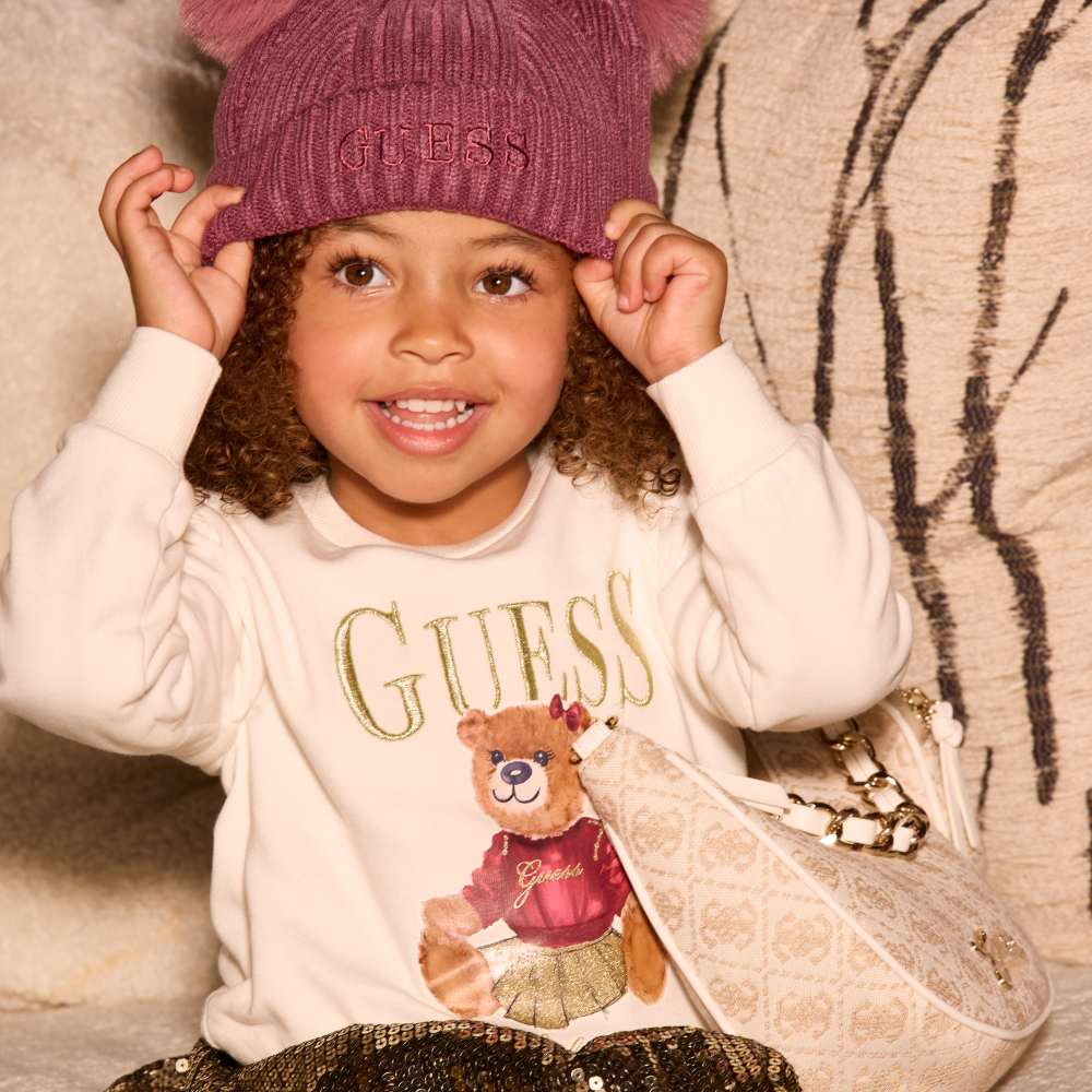 A smiling girl sat down wearing a pink bobble hat and Guess Kids top with a teddy on the front 