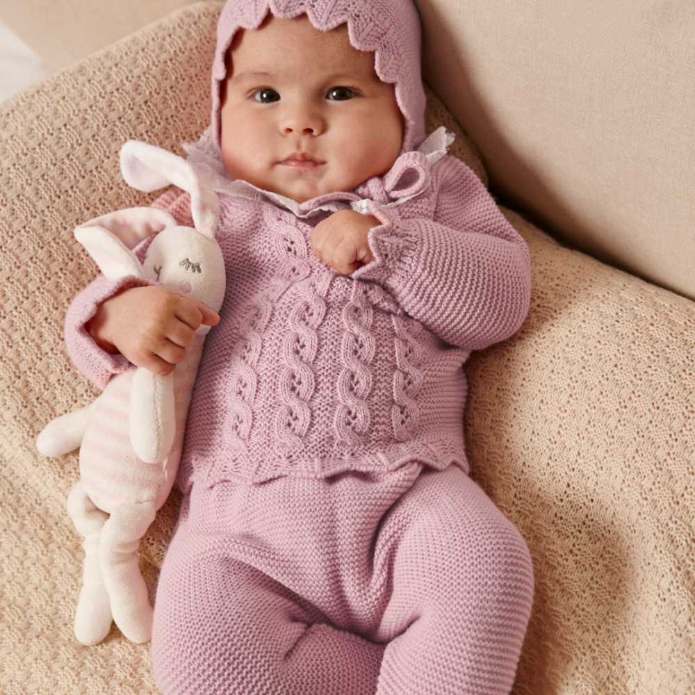 A baby lying down holding a teddy wearing a matching lilac knitted bonnet, jumper and trousers 