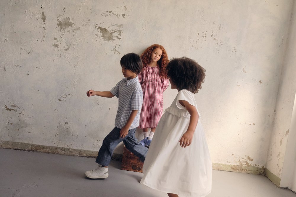Three children playing in a bare room wearing outfits by Kindred