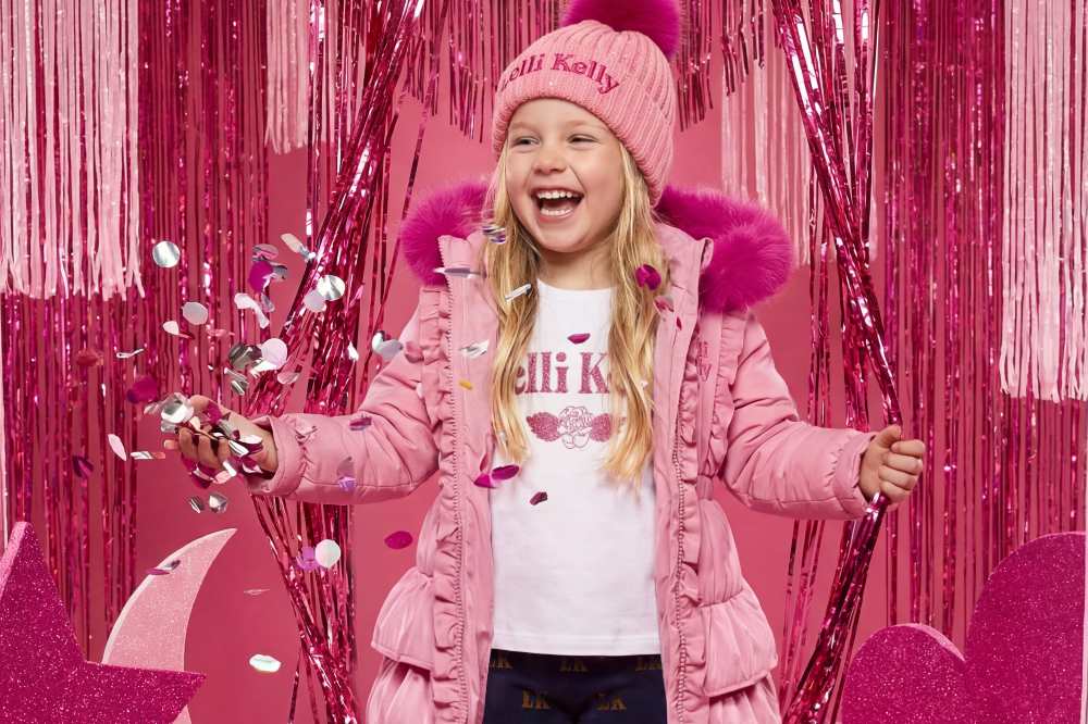 A girl in a pink winter coat and bobble hat by Lelli Kelly Clothing throwing glitter in the air in a pink room 