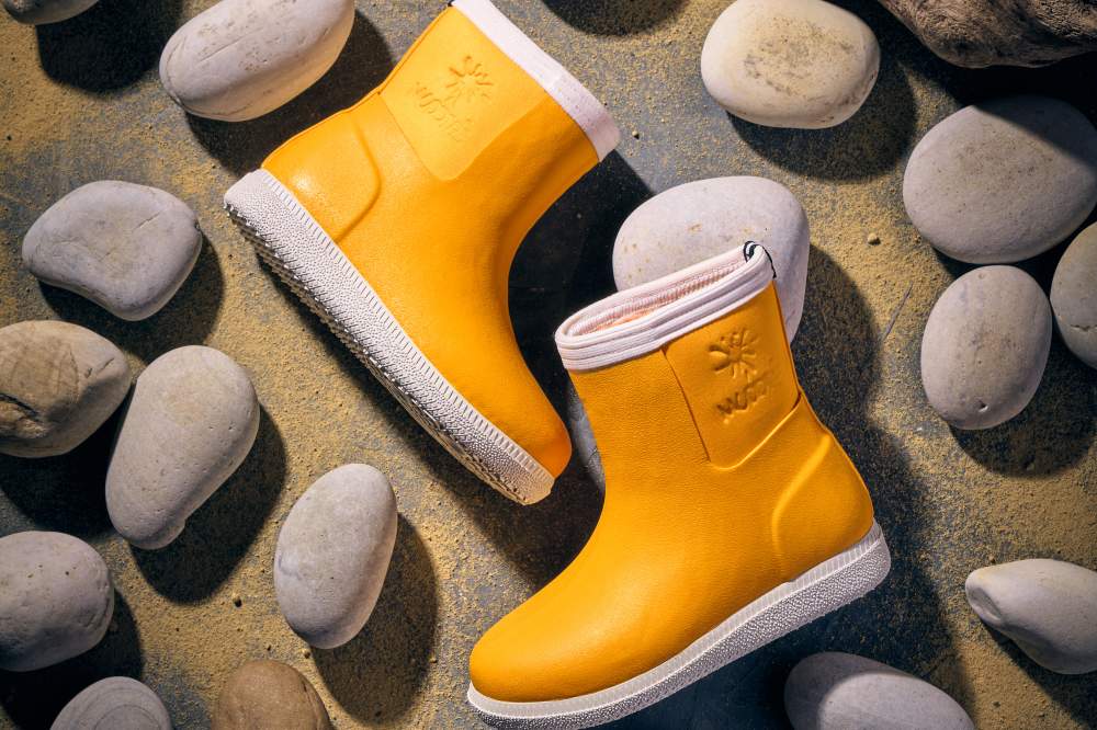 A pair of yellow barefoot outdoor children's boots by Muddies displayed on pebbles