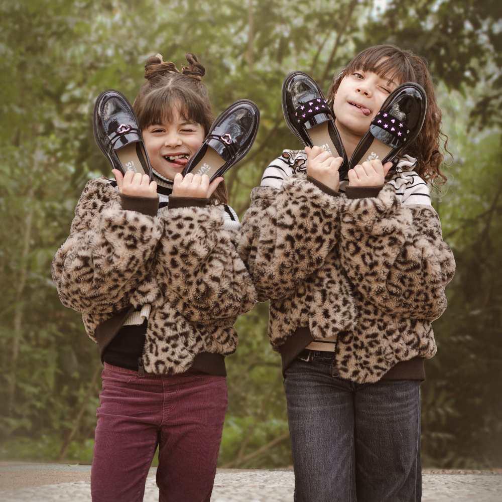 Two girls stood outside holding up pairs of black shoes 