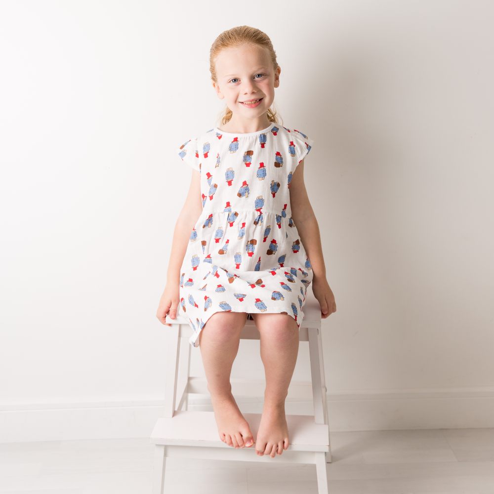A girl sat on a white chair wearing Paddington Bear print summer dress by Blade & Rose 