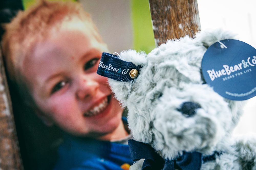 A pale blue bear by Blue Bear Co. shown beside a picture of a young boy