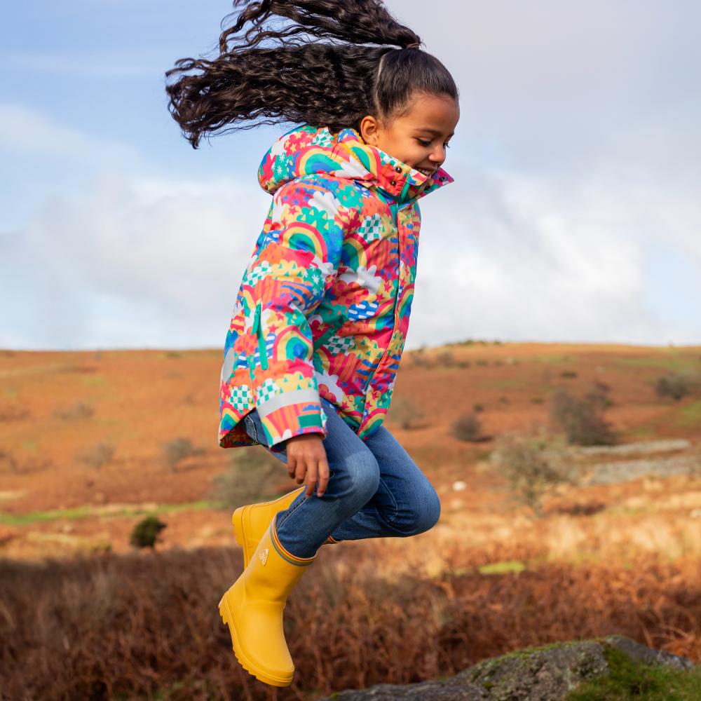 A girl outside jumping in the air wearing a brightly coloured winter jacket, jeans and yellow wellies 