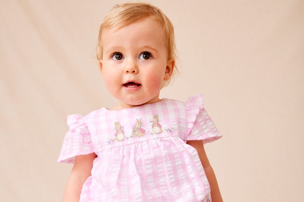 A baby wearing a pink gingham dress with Peter Rabbit embroidery by JoJo Maman Bebe