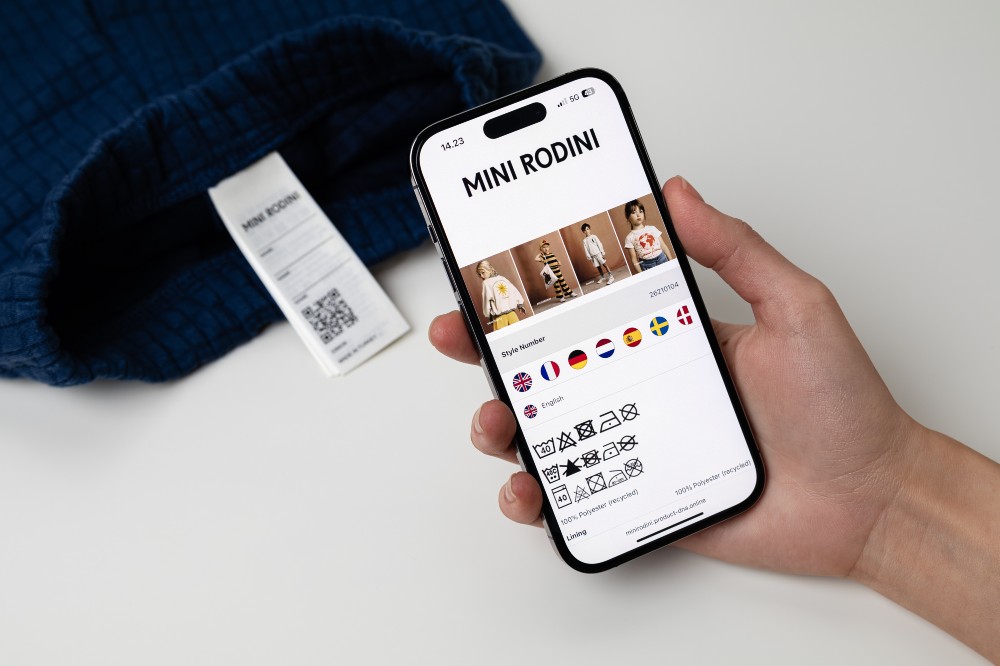 Someone's hand shown holding a smartphone in front of a Mini Rodini label garment