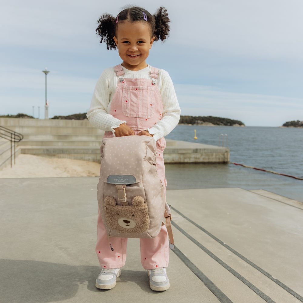 A girl stood outside by water in pink dunagrees holding a teddy bear rucksack 