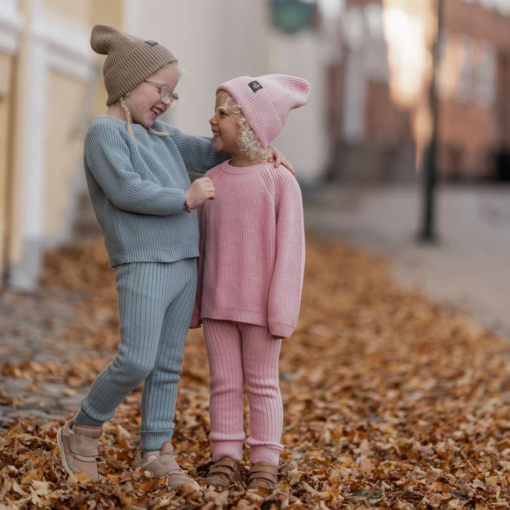 Two children in hats and winter outfits stood outside on a pavement covered in leaves 