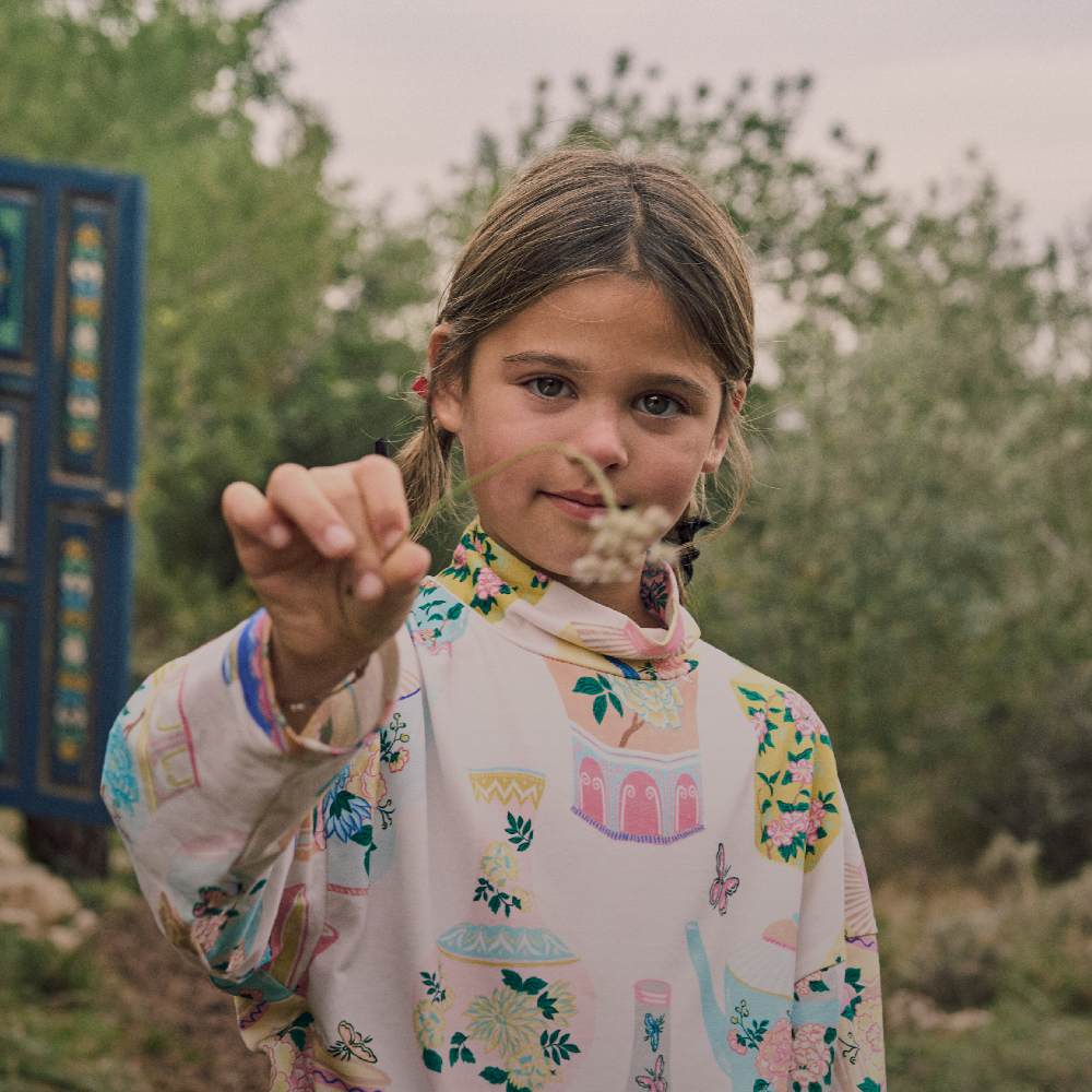 A girl stood outside in a printed top holding a flower stem to the camera 