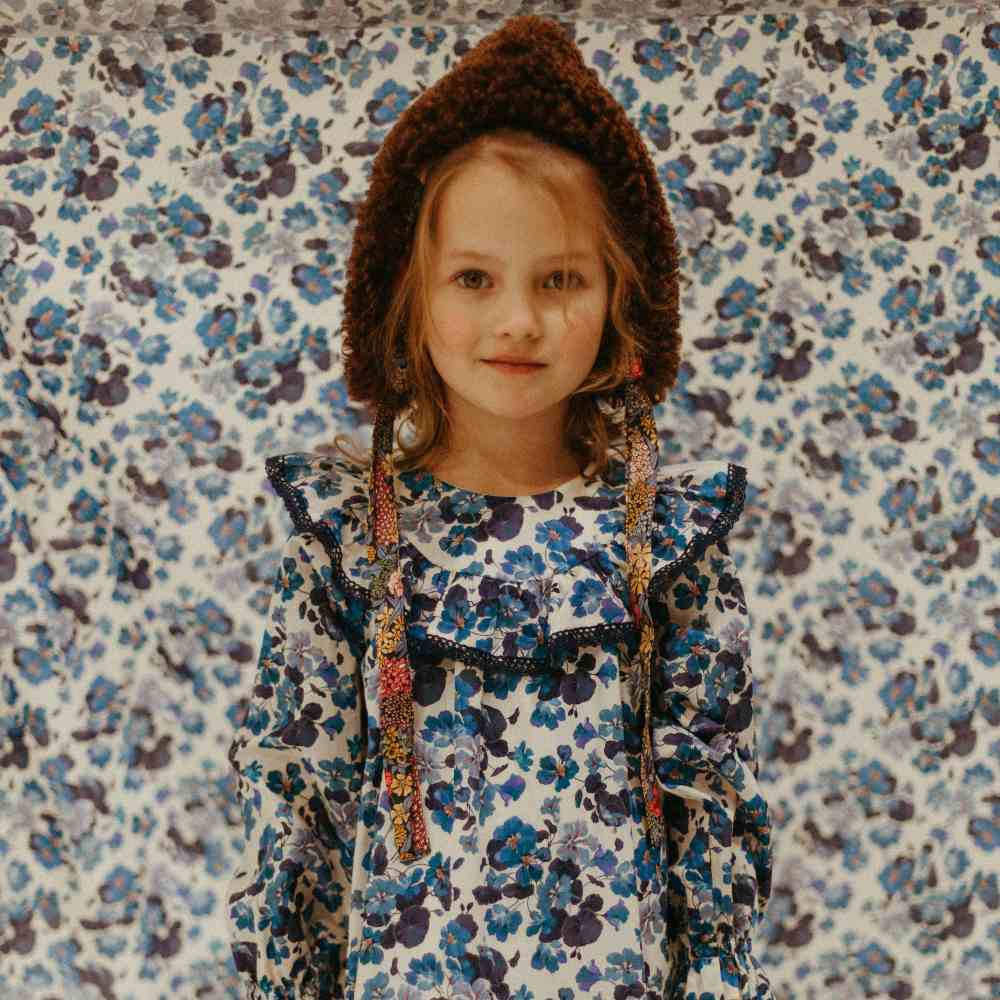 A girl in a hat and printed dress stood in front of matching printed wallpaper
