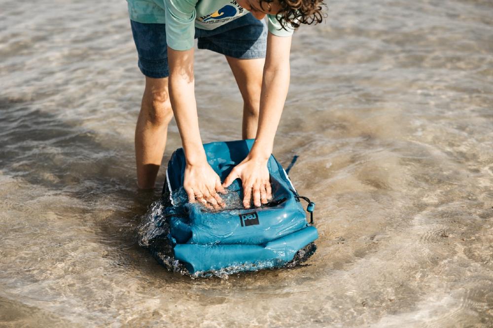 A child putting the Red Equipment Adventure Waterproof Backpack in the sea 
