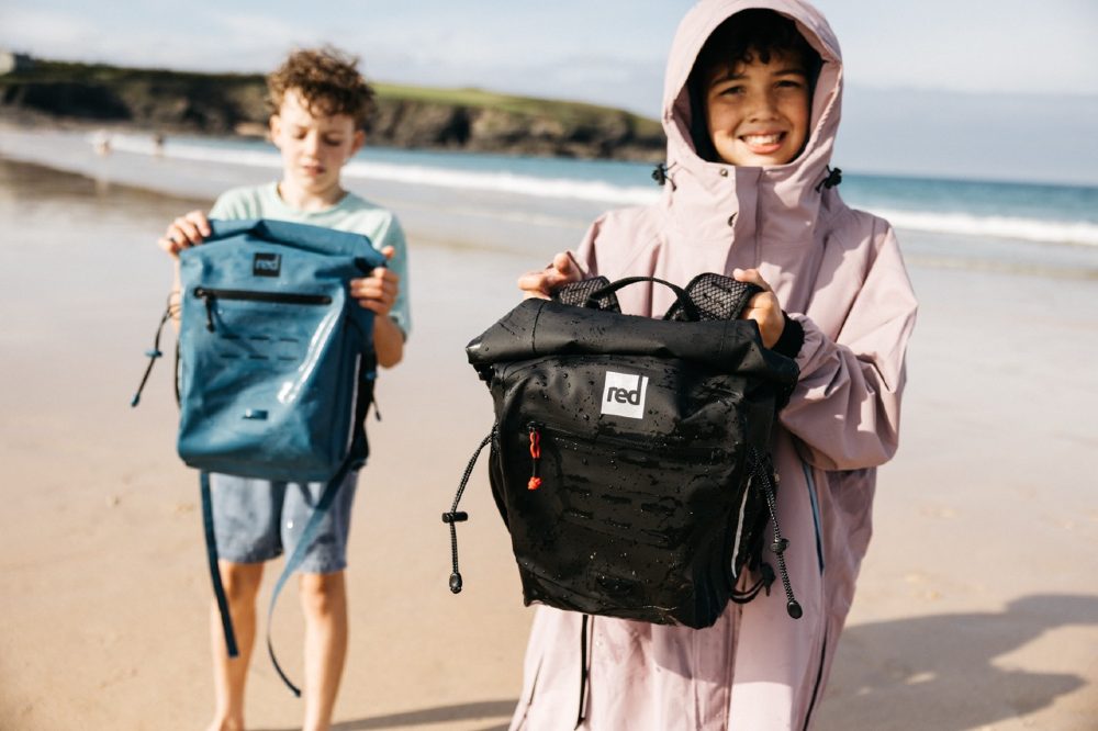 Two children on a beach holding up the new Red Equipment Adventure Waterproof Backpack