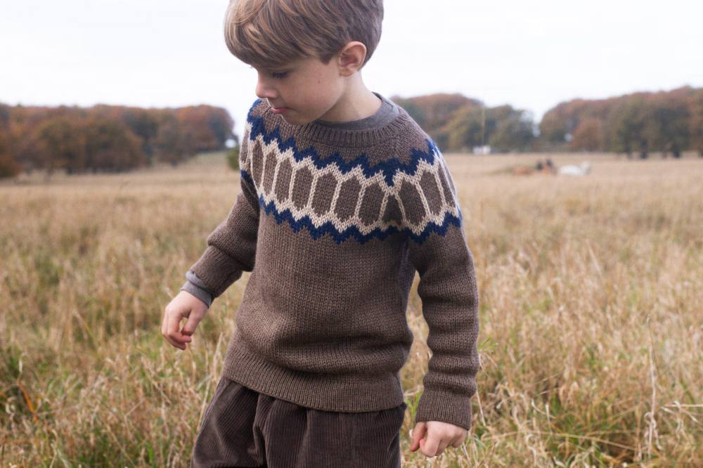 A boy in a knitted jumper and trousers walking through a field of tall grass