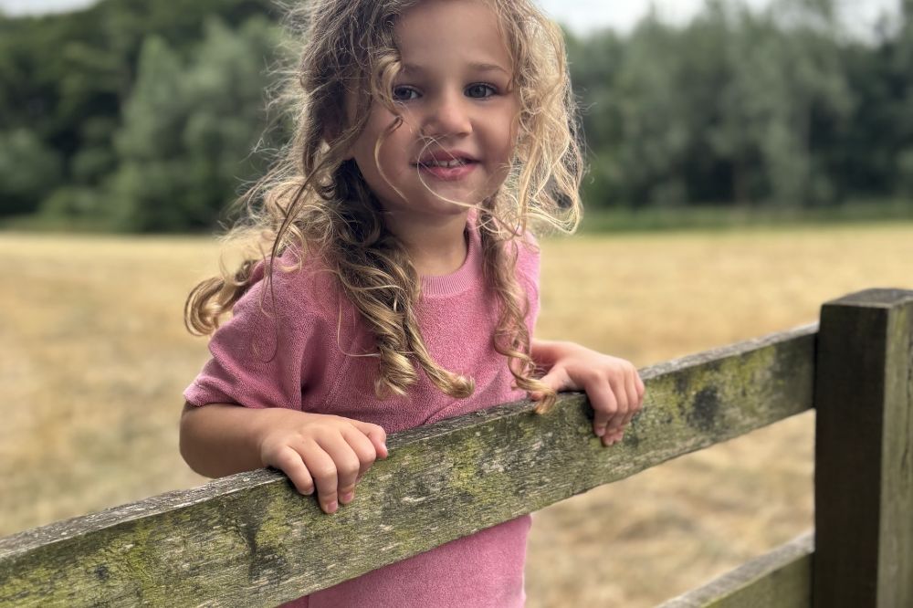 A girl stood on a fence in a field wearing a pink towelling T-shirt by Tots in Towelling