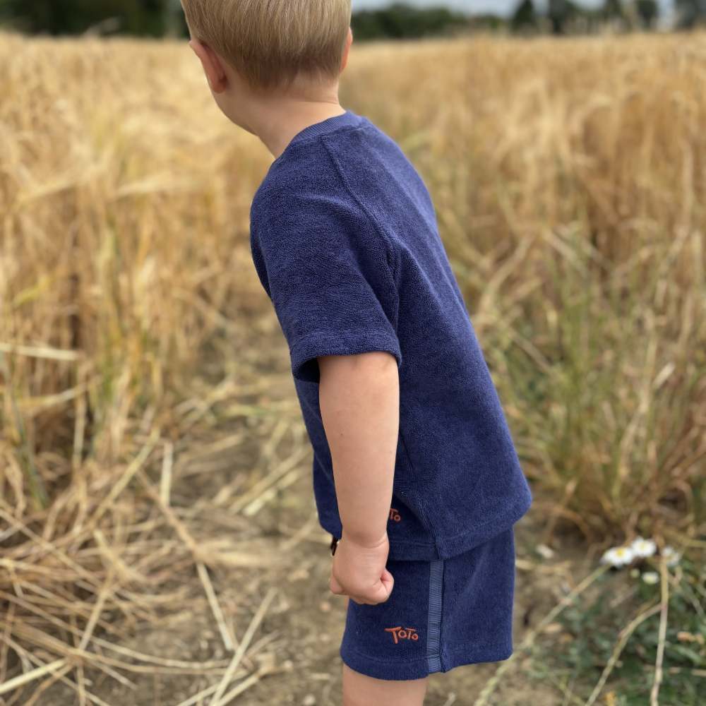A boy stood in a corn field wearing blue towelling shorts and T-shirt by Tots in Towelling 