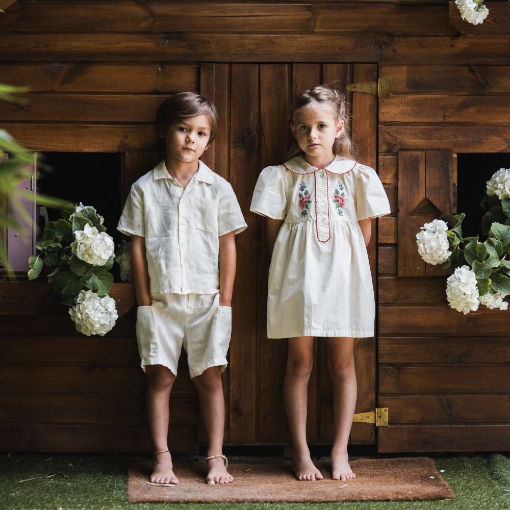 A boy and girl in white summer outfits stood in front of a wooden hut 