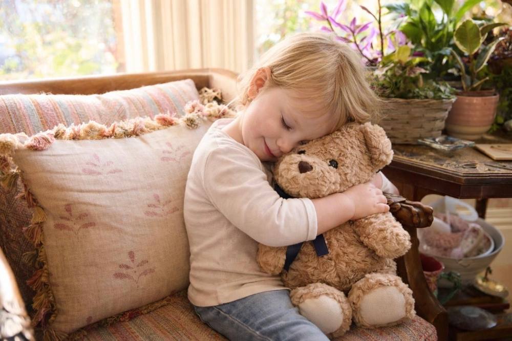 A young girl sat on a sofa hugging a teddy bear by Blue Bear Co.