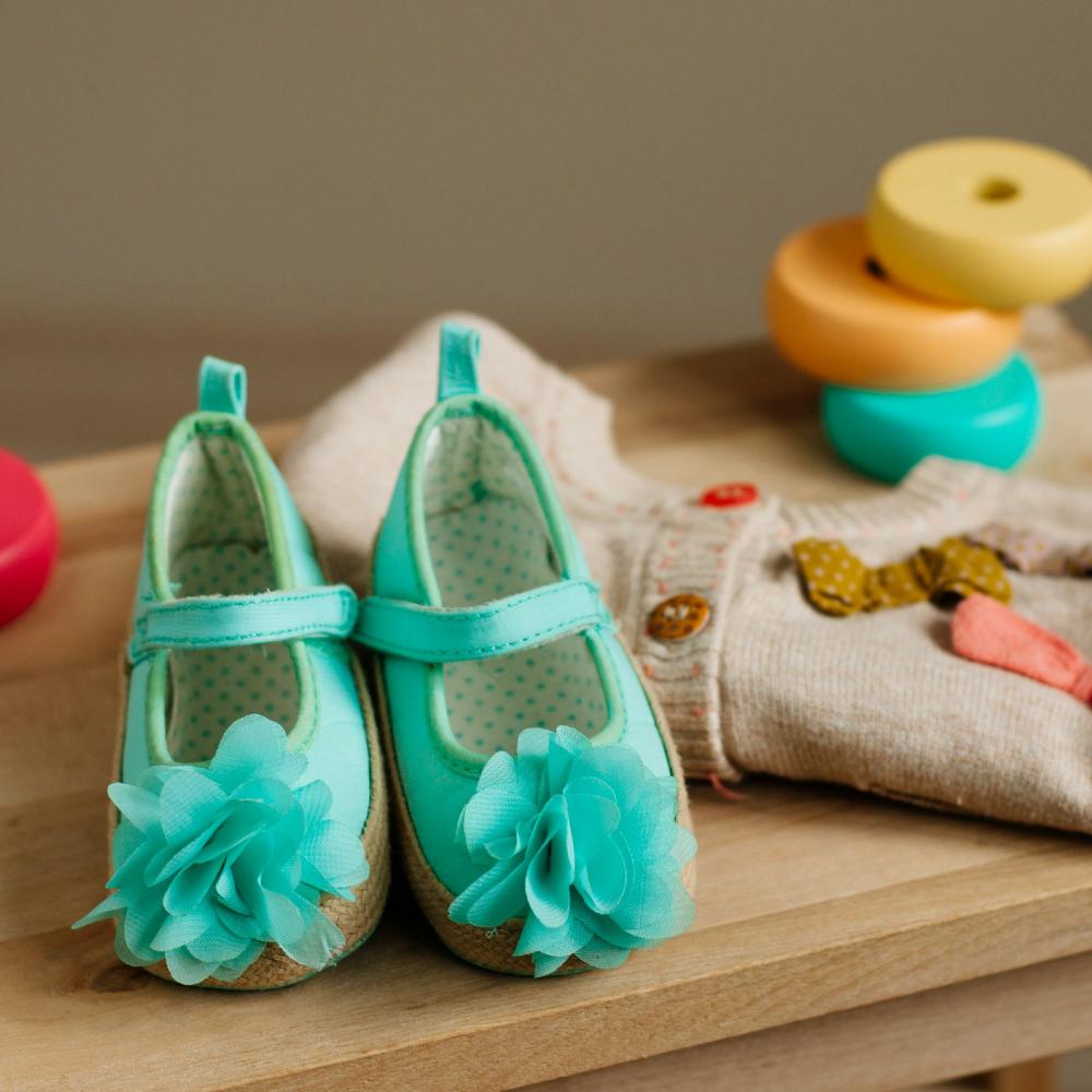 A pair of blue children's shoes with flower detail on the toes beside a children's cardigan and wooden toys 