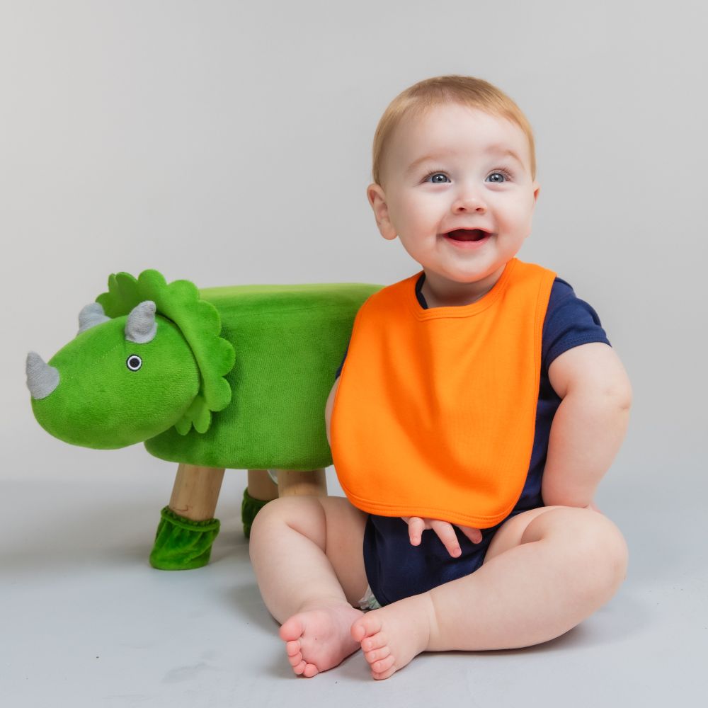 A baby in an orange bib sat beside a green dinosaur soft toy 