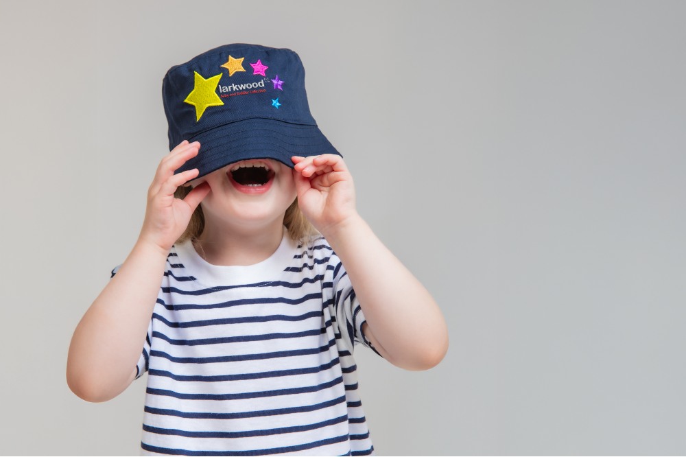 A laughing child in a striped T-shirt pulling a blue hat over their face with Larkwood and stars embroidered on it