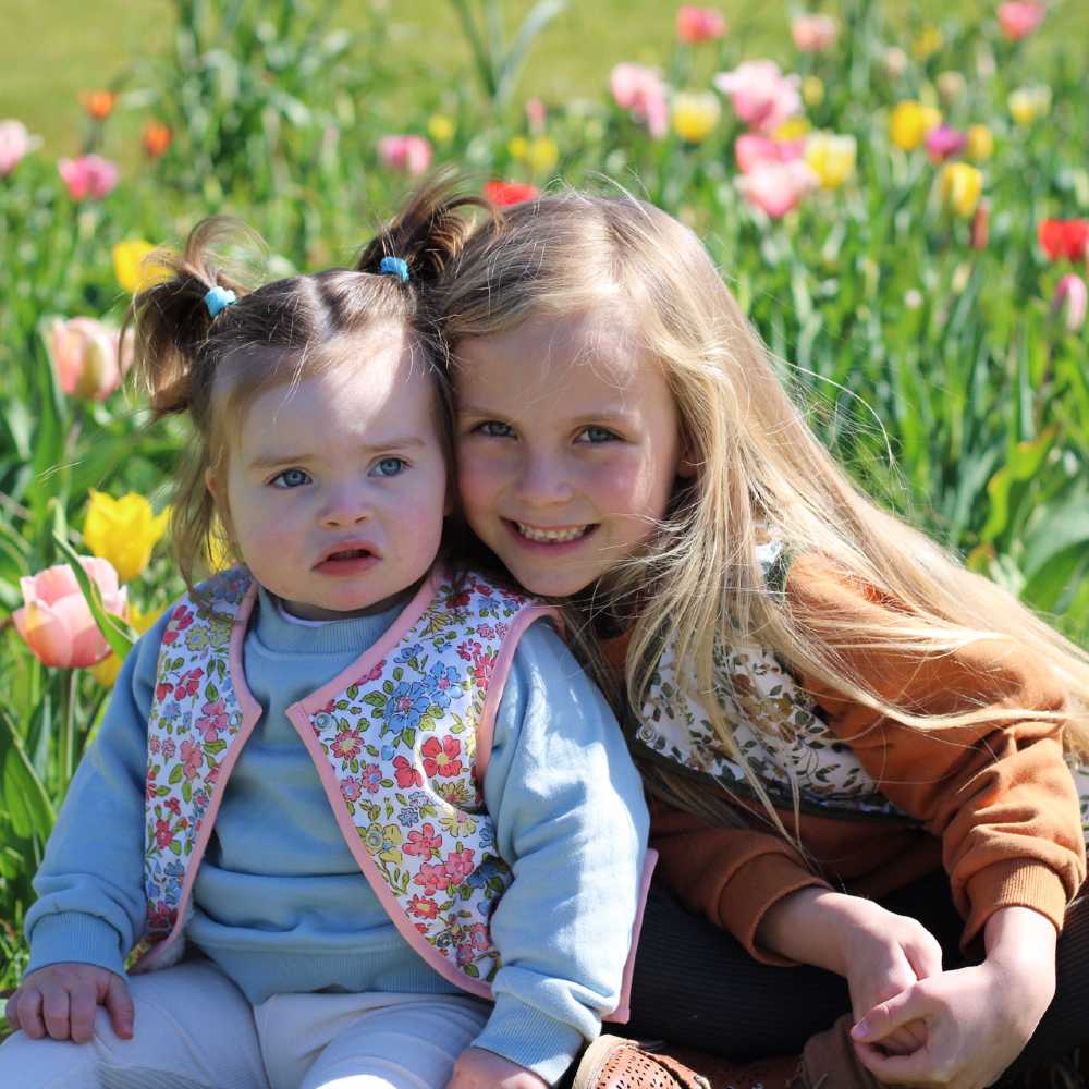 Two children wearing floral vest jackets by Little Scandi Baby sat in a field of flowers 