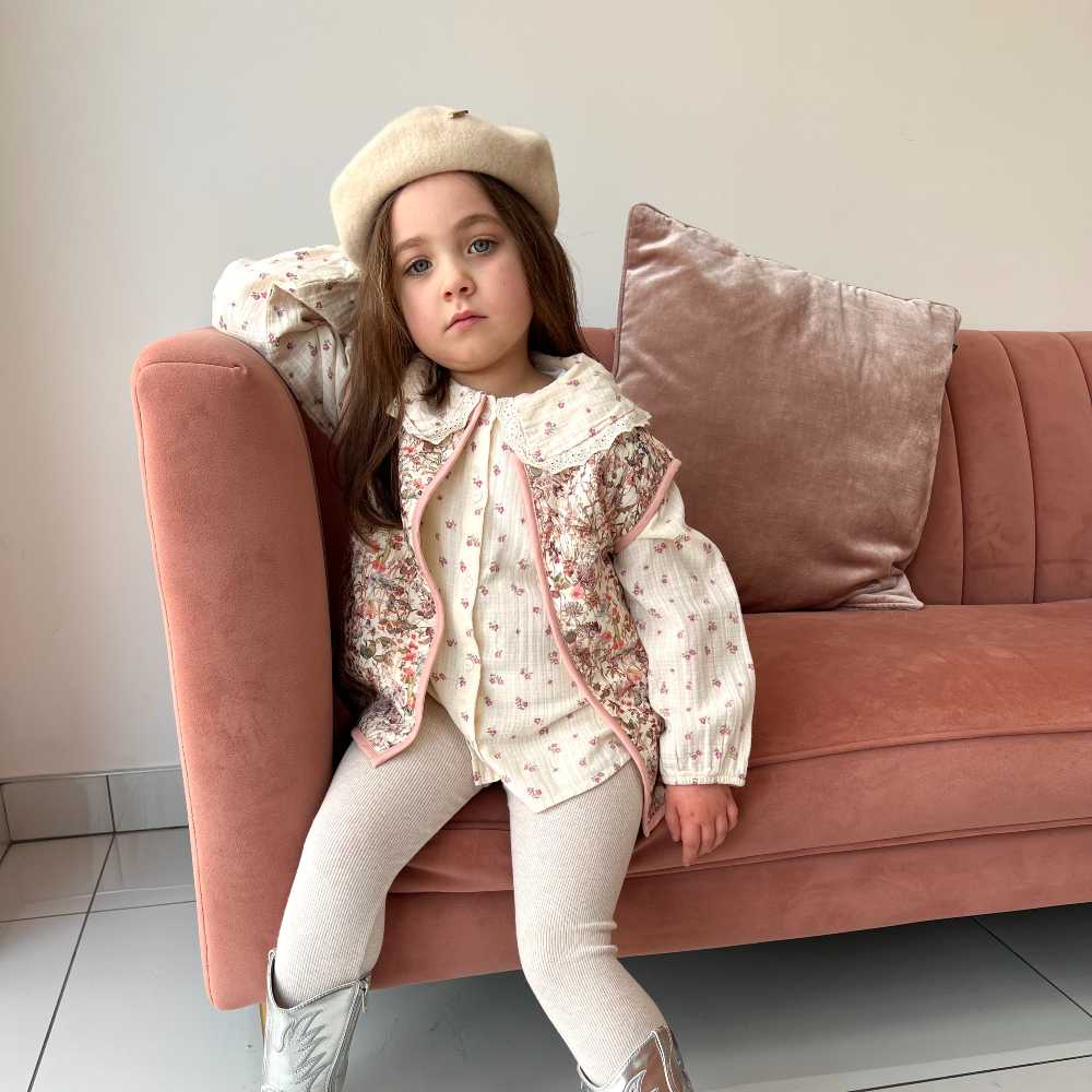 A girl sat on a pink sofa wearing a white beret, top and leggings and a floral vest by Little Scandi Baby 