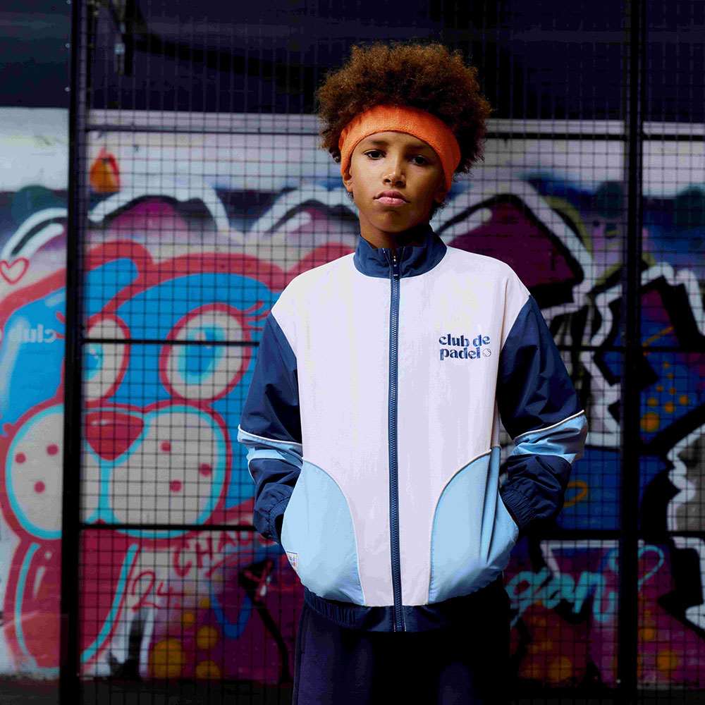 A boy stood in front a graffiti wall wearing a headband and bomber jacket from the M&S Kids x Club de Padel collab