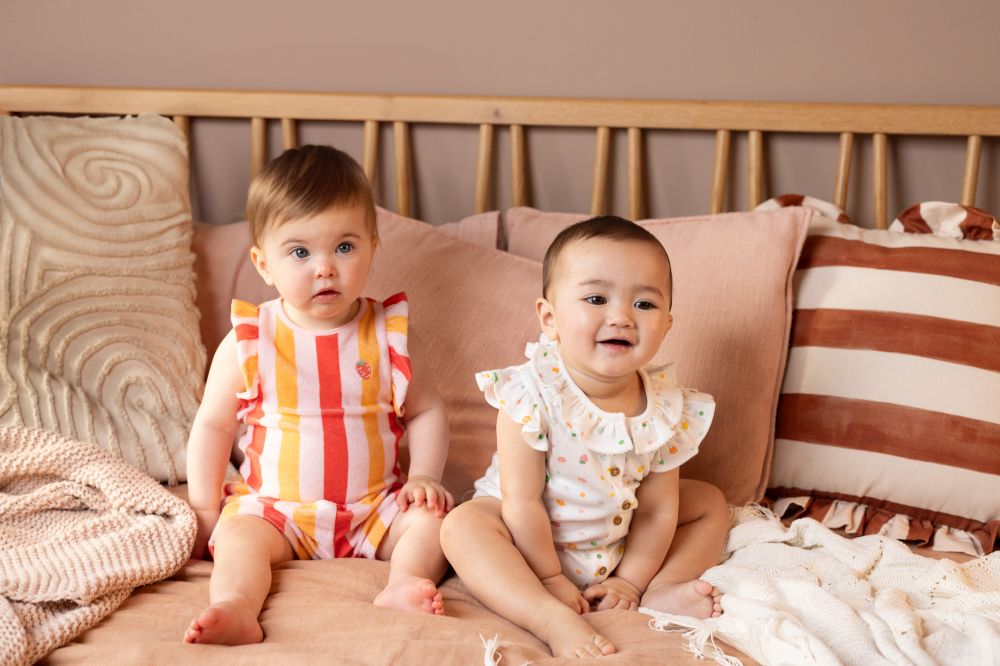 Two babies sat on a bed wearing summer outfits by Mini Cuddles 