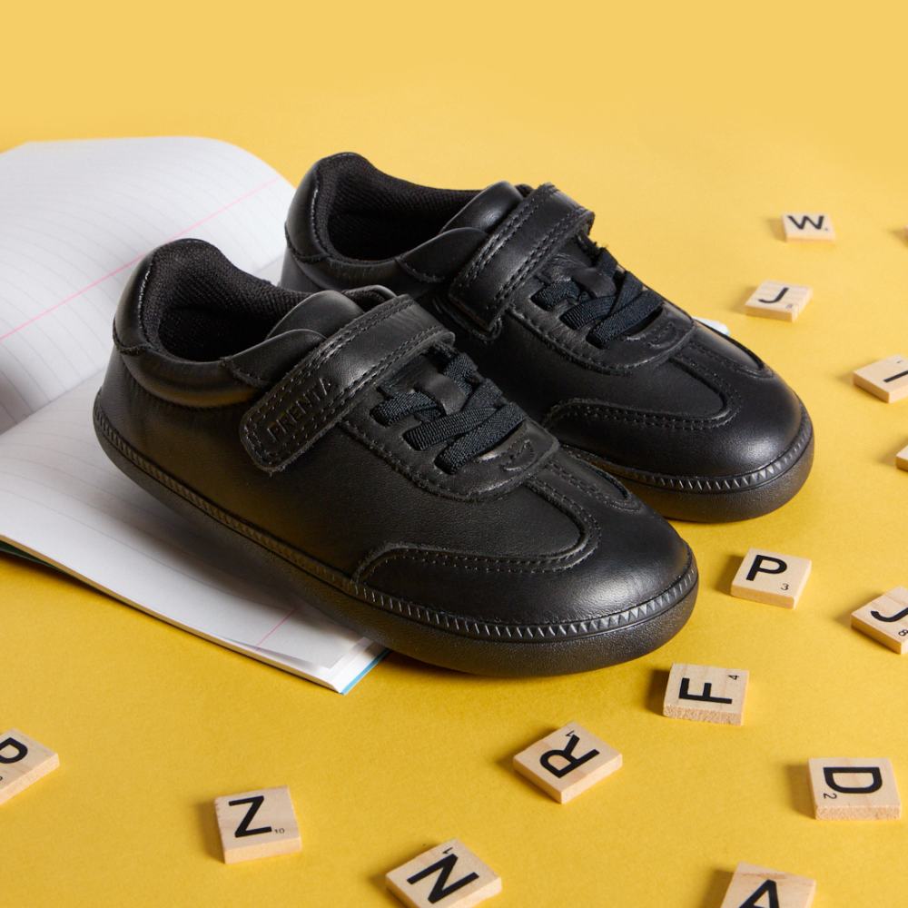A pair of black barefoot school shoes by PRENTA displayed on a yellow background with scrabble letters 