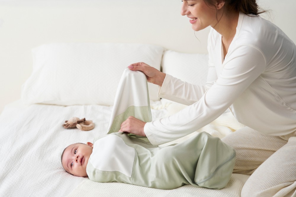 A woman wrapping a baby laid on a bed in a Momcozy blanket