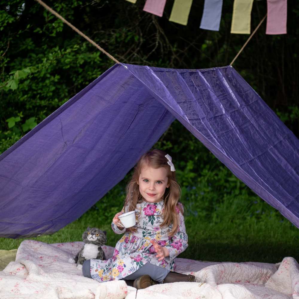 A girl outside sat beneath a purple den drinking from a cup 