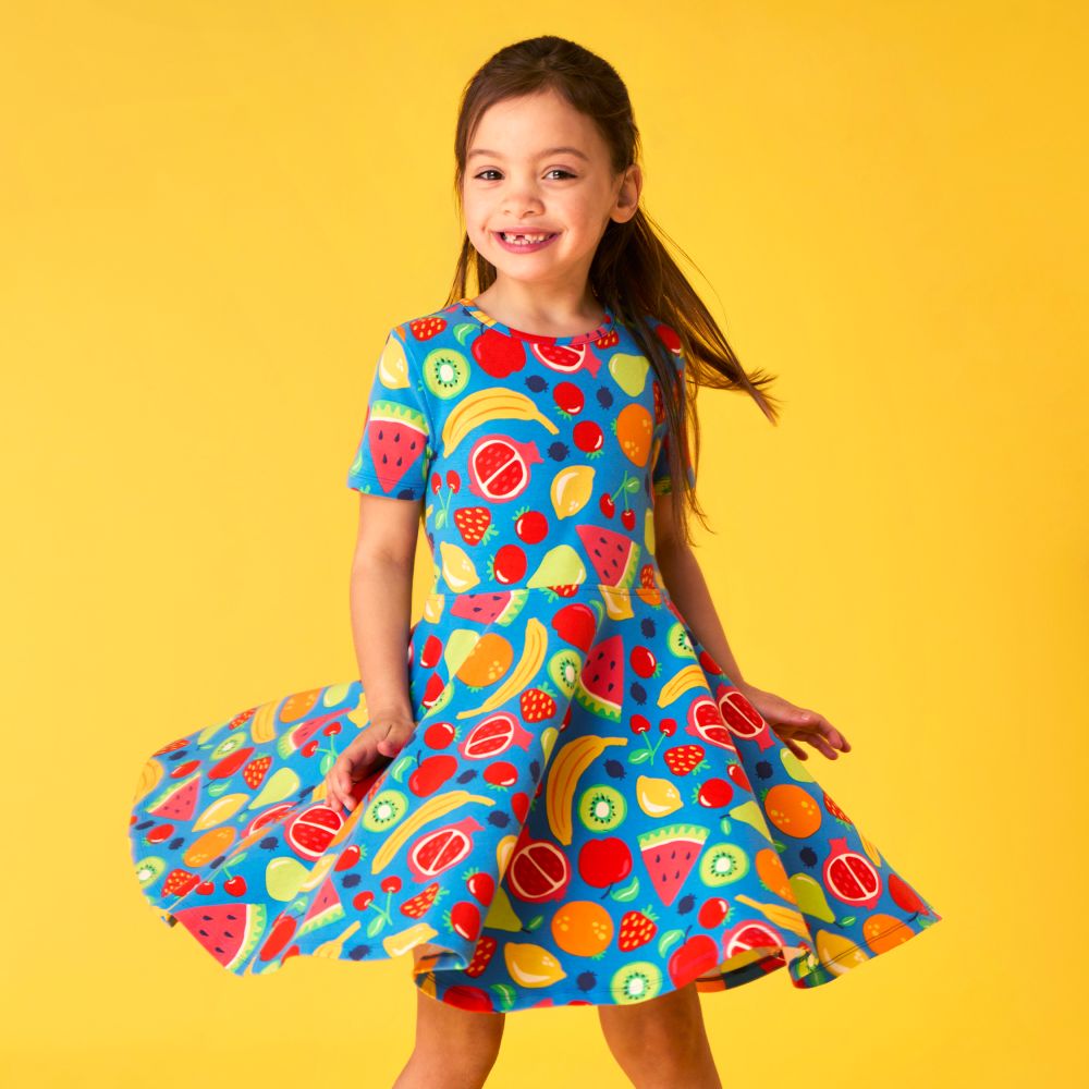 A smiling girl twirling in a fruit print dress by Frugi against a yellow background 