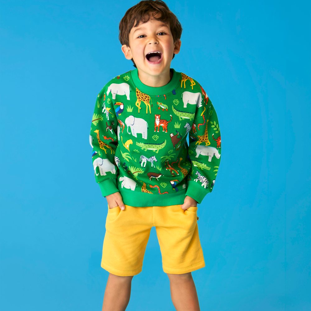 A smiling boy in a green top with animals on and yellow shorts by Frugi stood against a blue background 