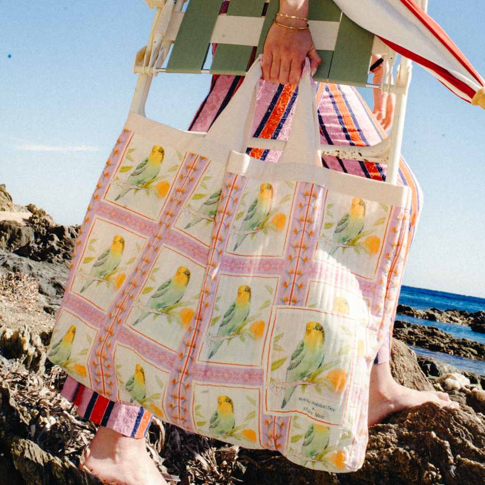 Someone walking on rocks on a beach carrying a large bag with a bird print from the Maison Mangostan x Studio Noos collaboration