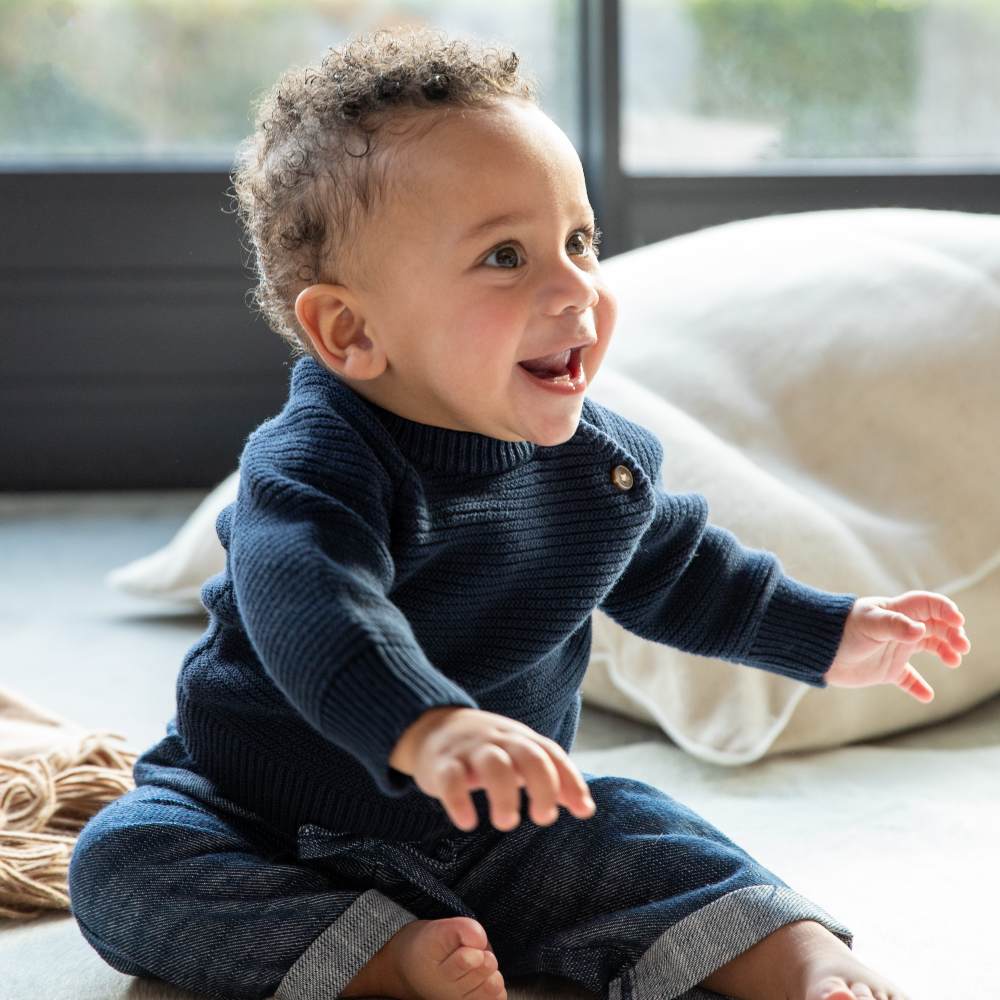 A smiling baby sat on the floor in a navy jumper and trousers 