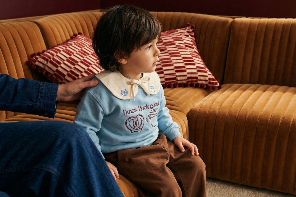 A young boy in a pale blue jumper and brown trousers sat beside a woman on a sofa