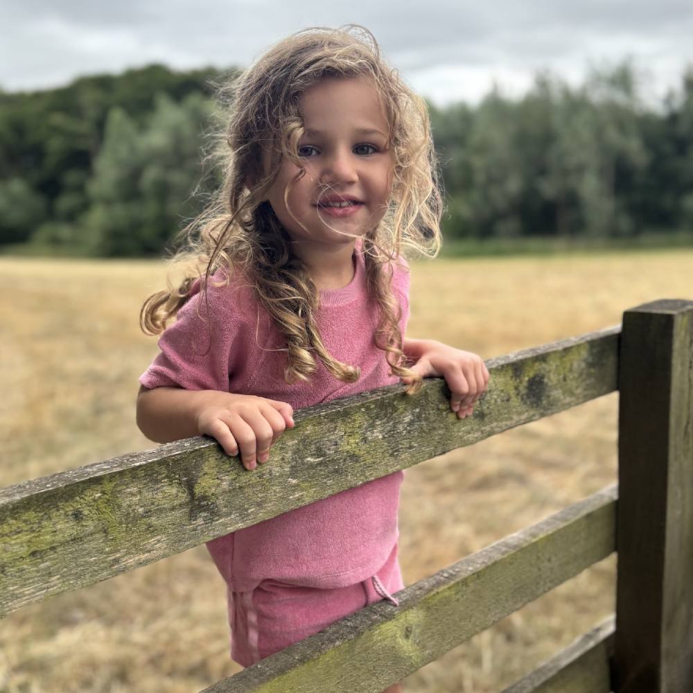 A girl in a pink towelling top and matching shorts stood on a fence in a field 
