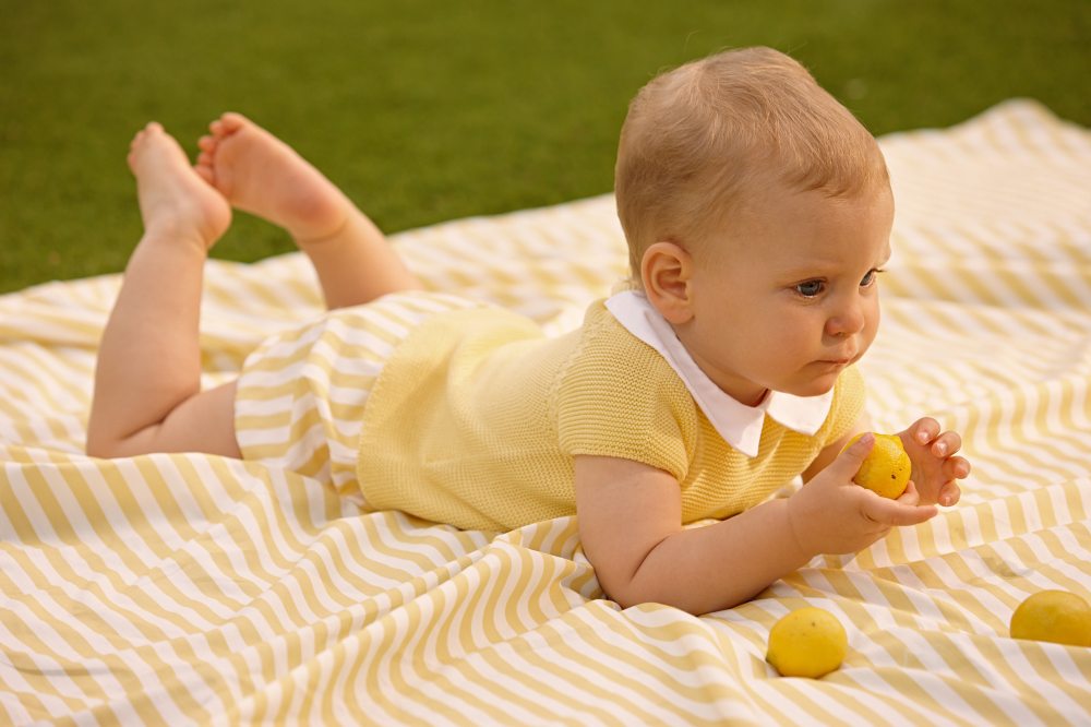 A baby ina yellow summer outfit lying on a blanket outside holding a lemon