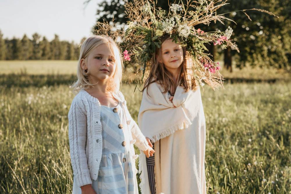 Two girls in summer outfits stood in a field with one wearing a crown of flowers