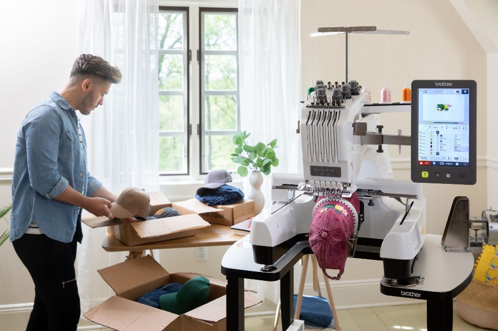 A man stood holding a baseball cap next to a Brother embroidery machine