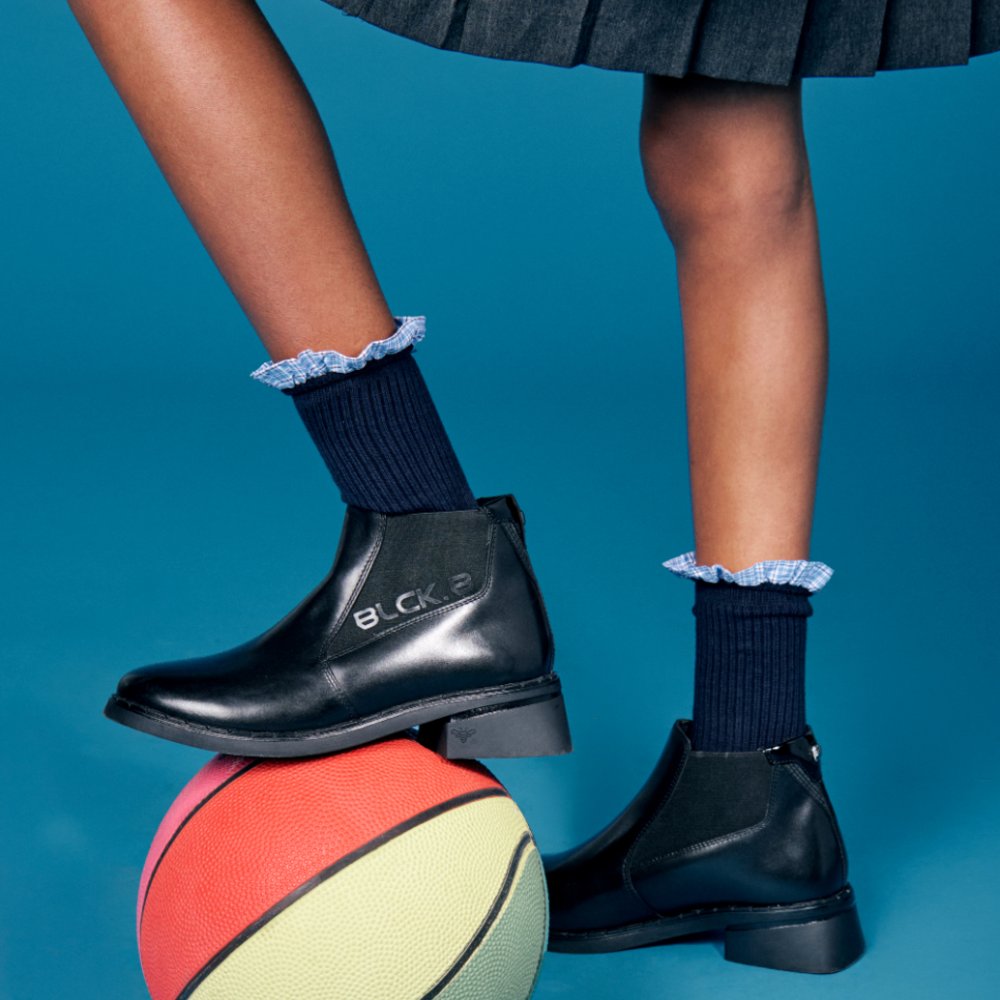 A child's feet shown wearing black school boots by BLCK.B with one foot resting on a ball