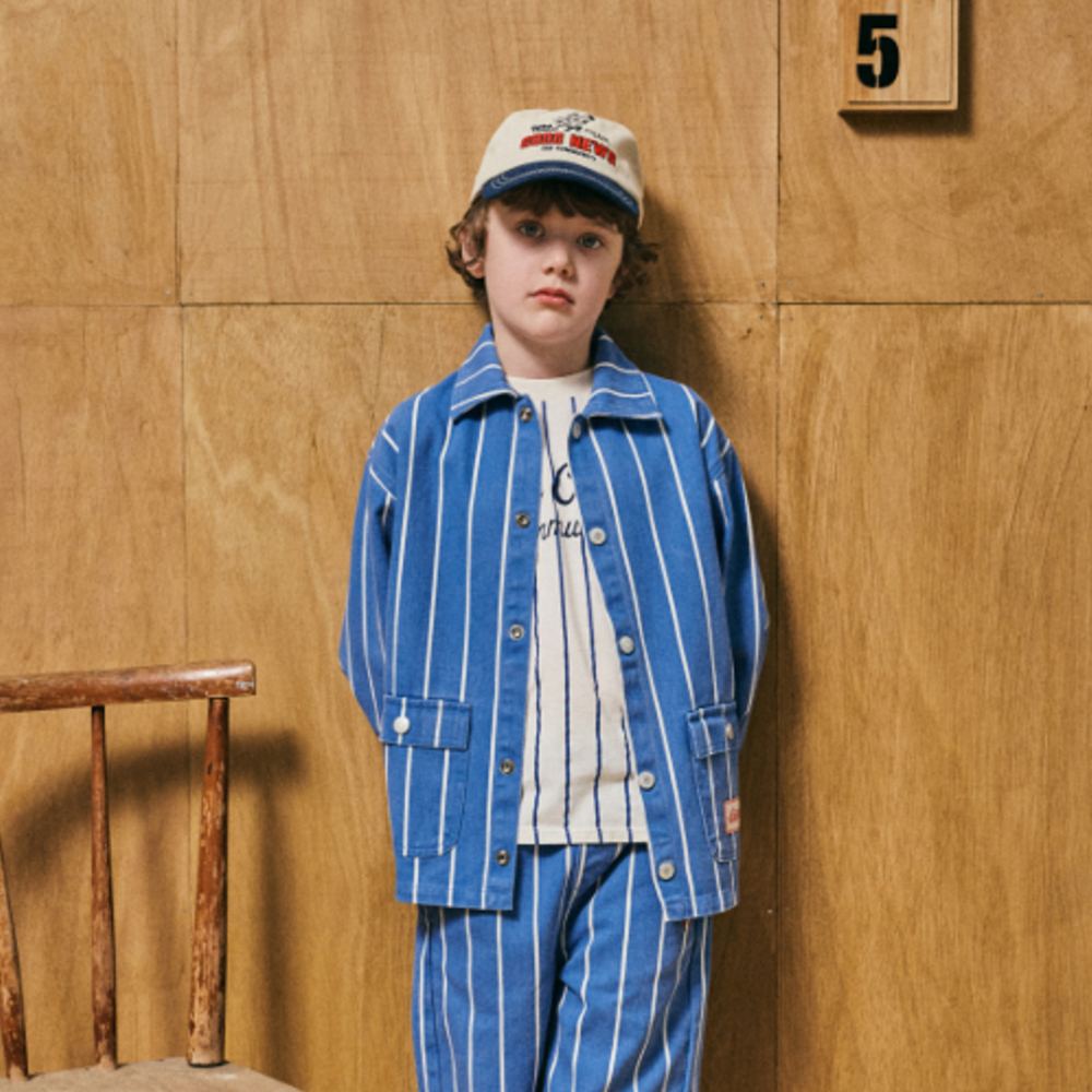 A boy in a blue striped shirt and trousers and a cap stood against a wooden panelled wall