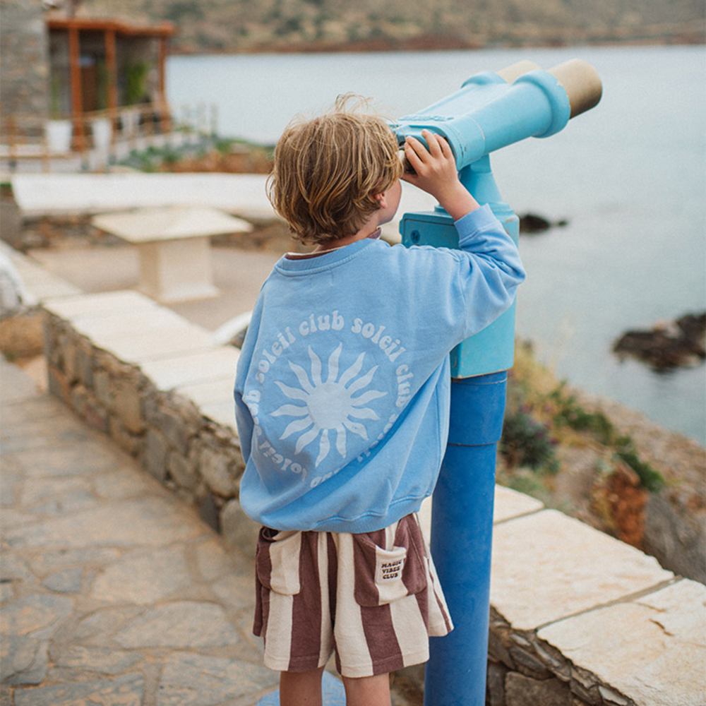 A boy in a blue sweatshirt and striped shorts looking through a telescope out to sea