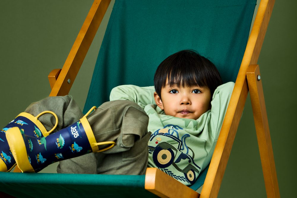 A boy sat in a green deckchair wearing a top, trousers and wellington boots from the JoJo Maman Bebe Transitional collection