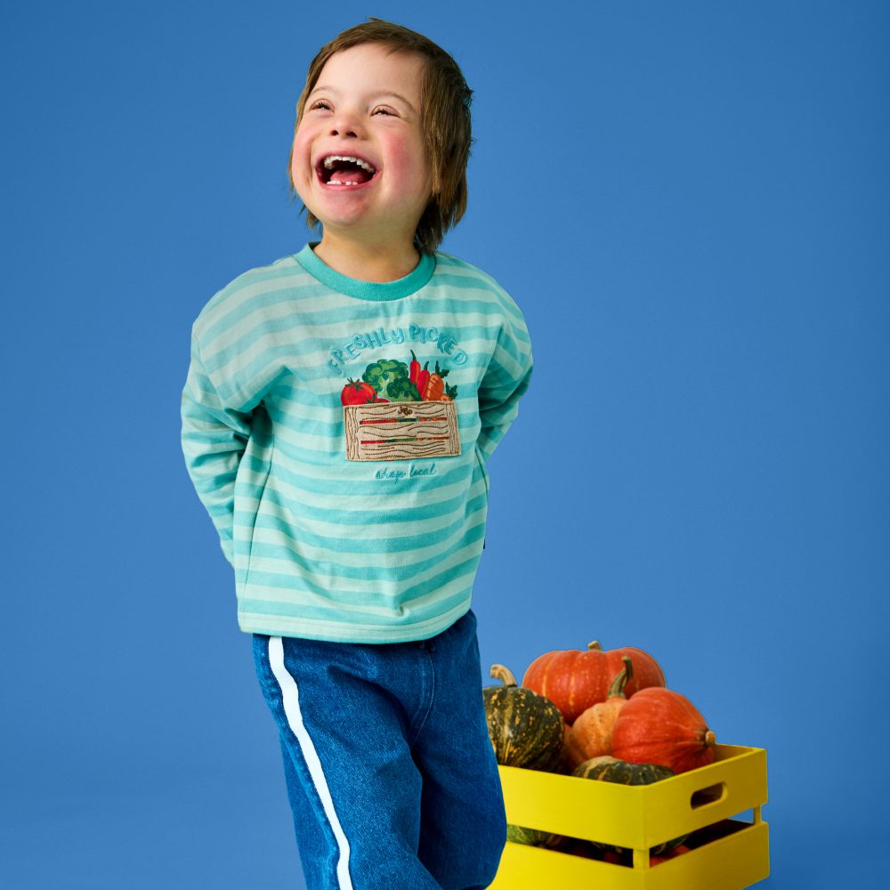 A smiling boy in blue trousers and a blue top with a vegetable applique stood next to a box of vegetables 