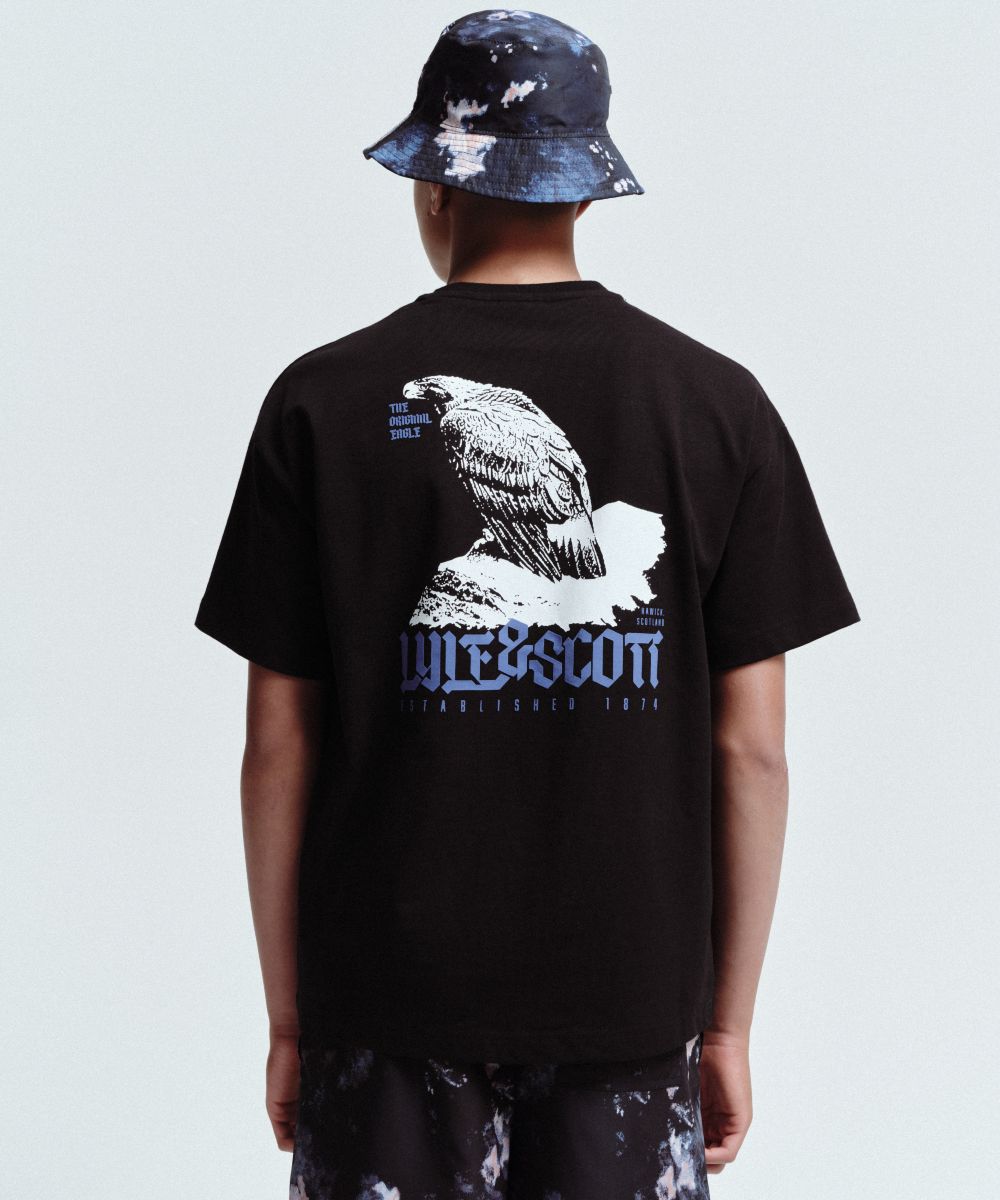 A boy with his back to the camera wearing a hat, graphic T-shirt and trousers by Lyle & Scott Kidswear