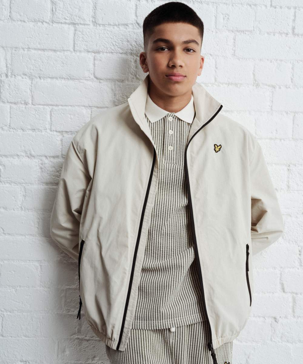 A boy stood against a white wall wearing a jacket, polo and trouser ser by Lyle & Scott Kidswear